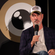 Roland Emmerich, in a white tee, blue jacket, and hat labeled "Deus," sits in front of a backboard with a stylized camera lens logo on it.